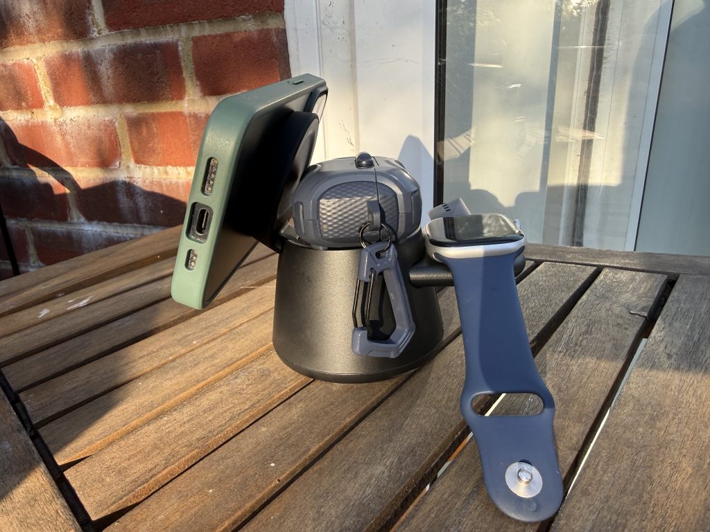 Close-up of a compact grey Belkin electronic charging device with a green iphone and blue iwatch on a sunlit wooden table, against a brick wall; quiet, practical mood.