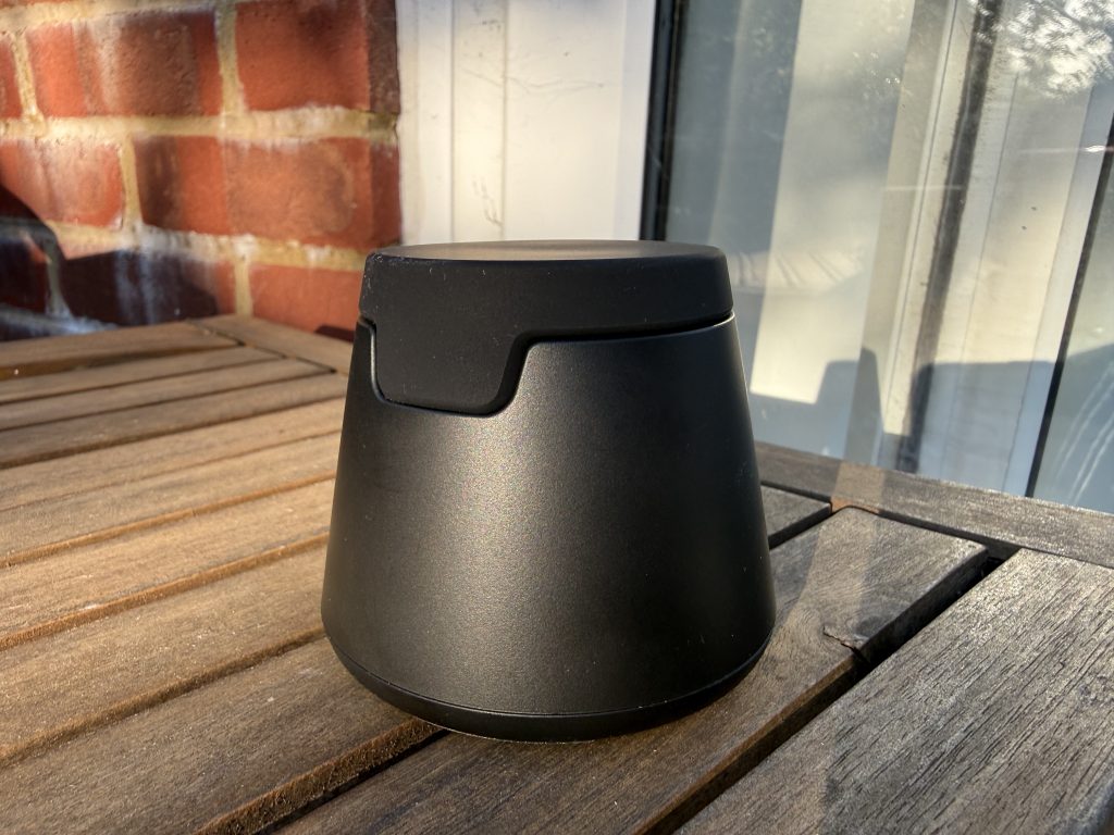 Matte black, rounded Belkin modular charger with a fitted lid sits on a wooden patio table in sunlight, against a brick wall and glass door.