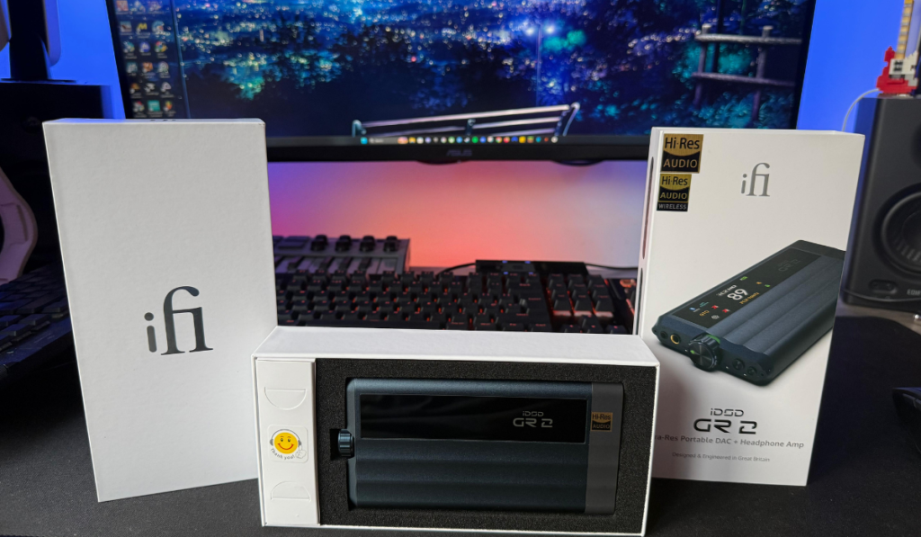 iFi iDSD GR 2 portable USB DAC and headphone amplifier displayed with its box on a desk, against a glowing cityscape monitor and studio speakers.