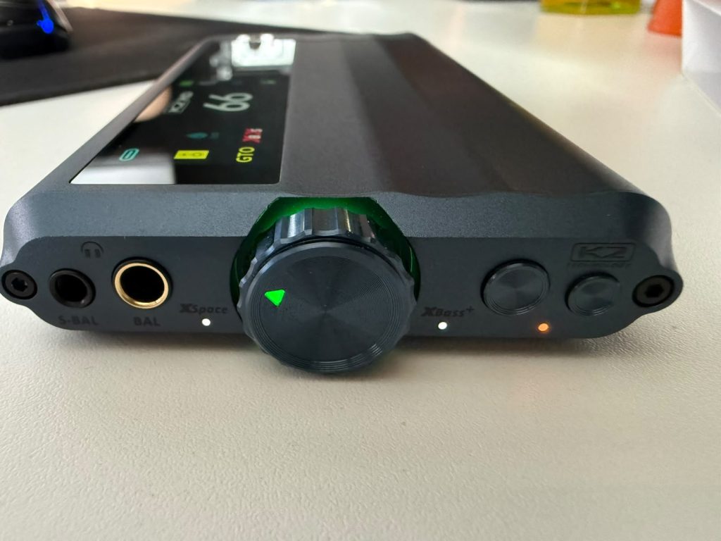 Close-up of a black portable headphone amplifier on a desk, showing a large volume knob, headphone jacks, buttons, and green and orange lights.