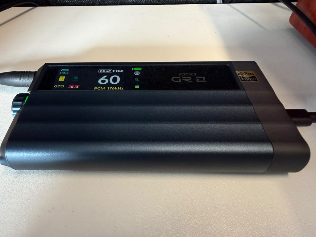 Close-up of a the iFi iDSD GR 2 with the touchscreen displaying the volume as “60” along with the audio input and output settings. It is resting on a white desk with cables attached, conveying a practical tech setup.