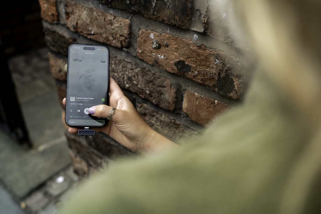 A person holds a smartphone displaying a music player beside a weathered brick wall, with a blurred green jacket in the foreground; candid urban mood. The iFi UP Play Max is inserted into the bottom of the audio device.