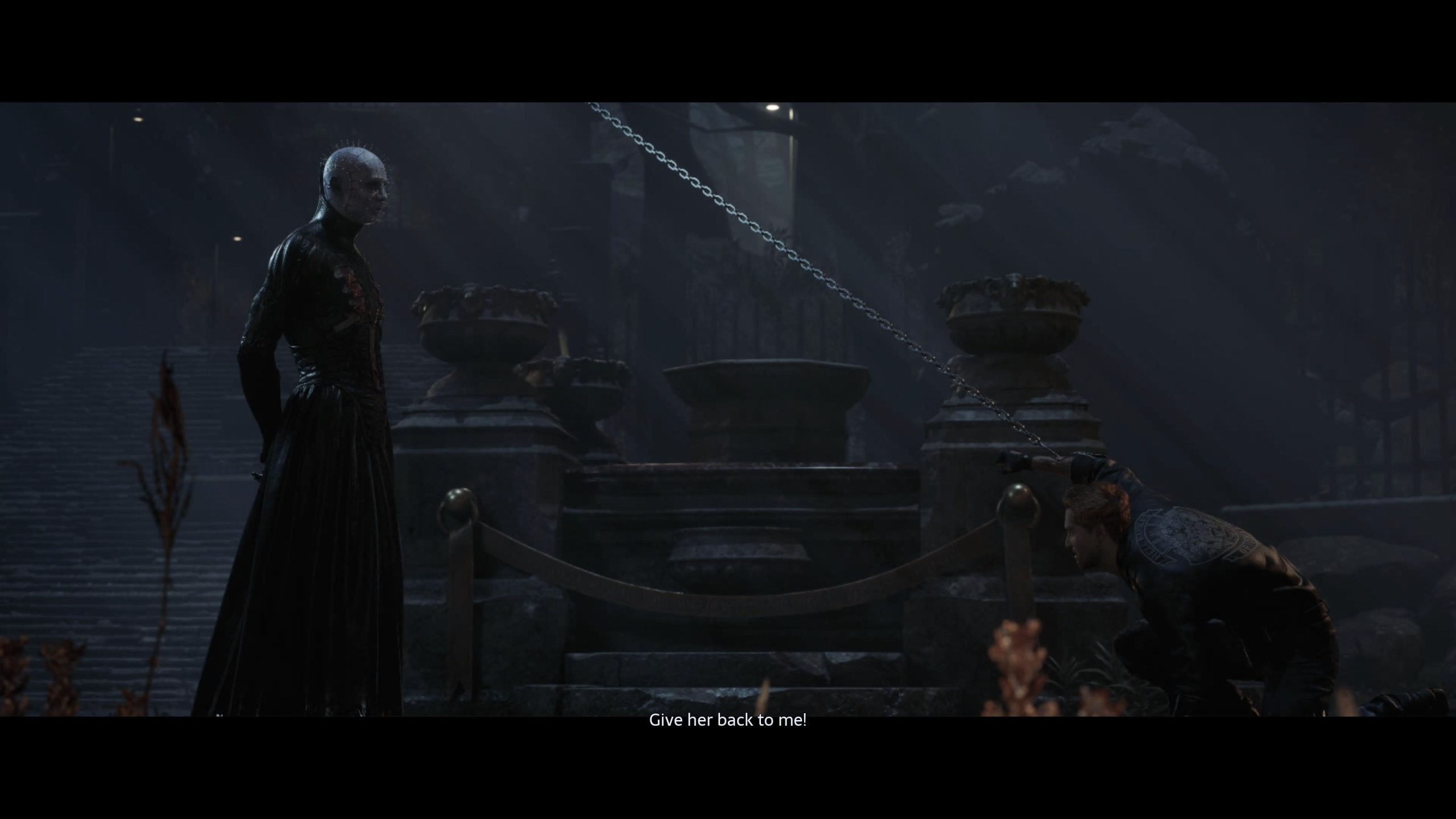 Pinhead and Aiden are in a graveyard. Pinhead is on the left whilst Aiden is crouched on the right hand side. There is a chain through Aiden's arm, holding it up